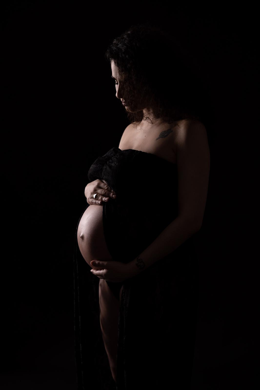 Futura mamma di profilo con le mani sul pancione, ritratto maternity in luce soffusa